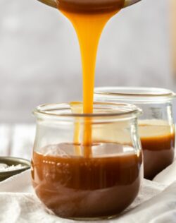 Caramel sauce being poured from a saucepan into a glass jar. Another sits behind