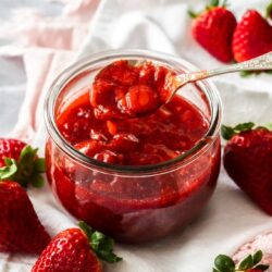 A spoon digging into strawberry topping in a round glass jar on a white napkin with strawberries around it