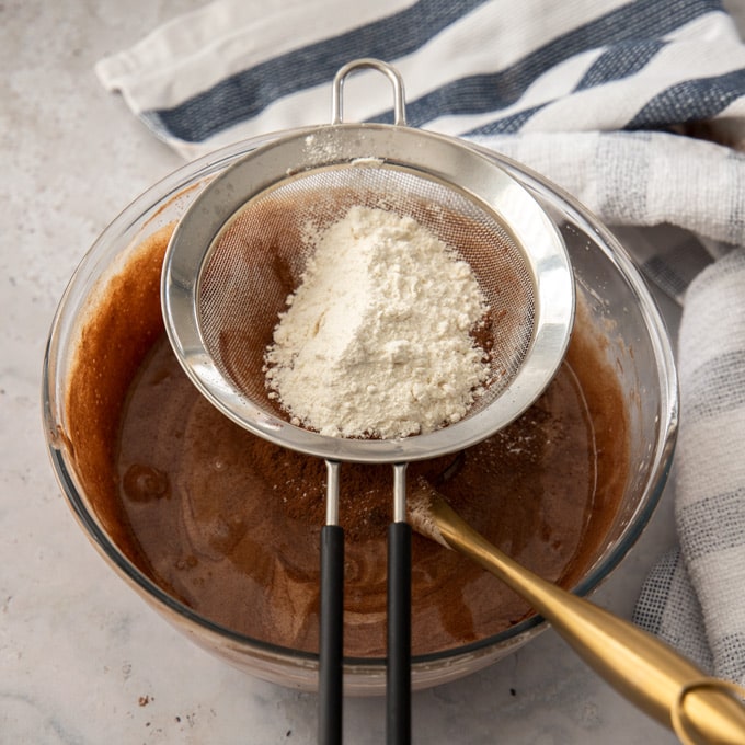 A sieve with flour and cocoa sitting over a glass bowl of chocolate batter