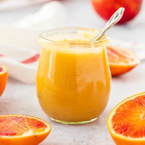 A glass jar filled with orange curd with a spoon sticking out.