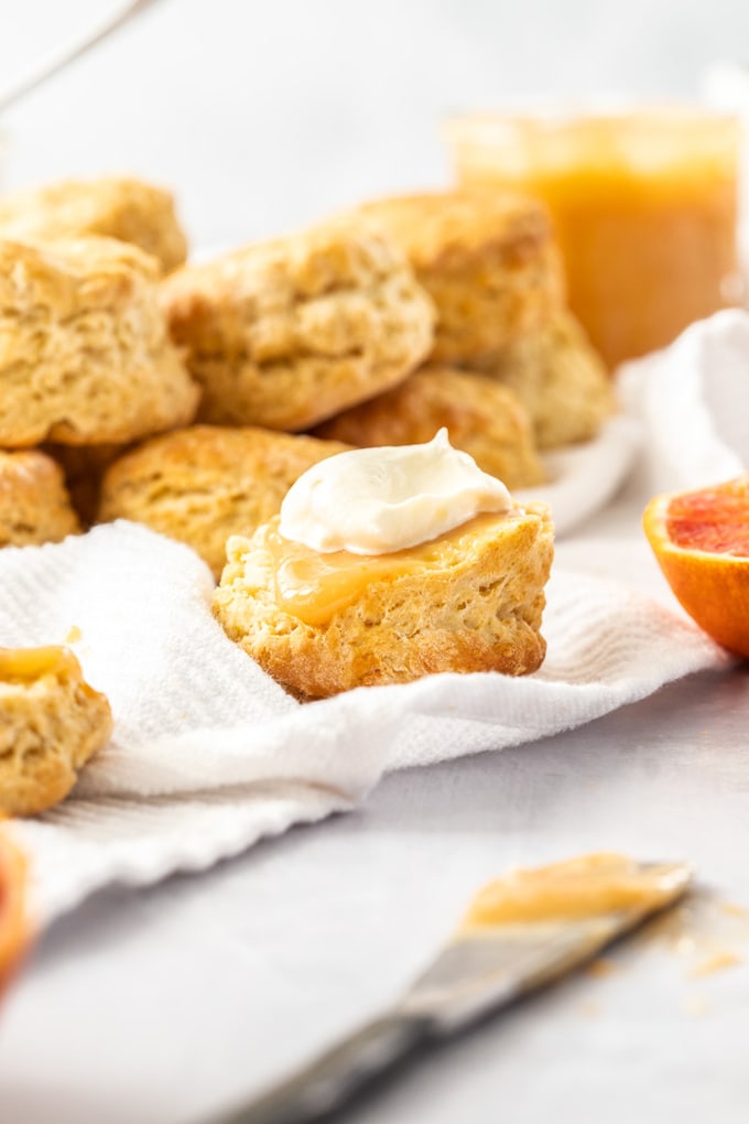 Half a scone topped with orange curd and cream sitting on a white tea towel. A pile of scones sits behind it