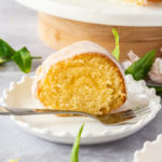 A slice of vanilla bundt cake on a white plate with a fork in front. Some lemon tree leaves scattered around