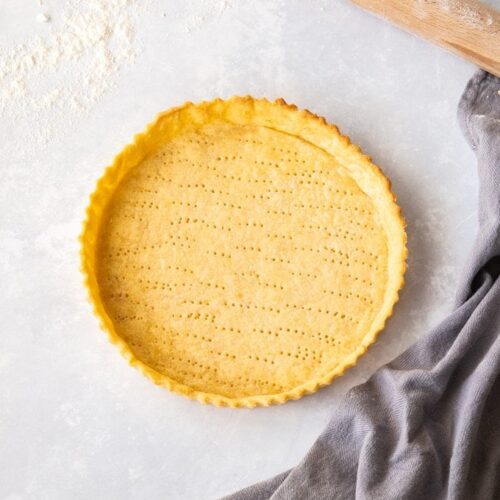 Birdseye view of a pastry tart shell on a floured worktop with a grey tea towel and rolling pin nearby.