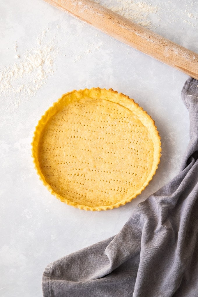 Birdseye view of a pastry tart shell on a floured worktop with a grey tea towel and rolling pin nearby.