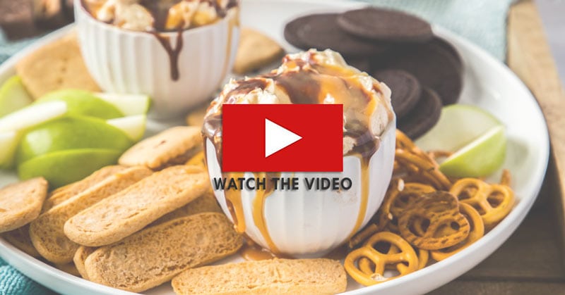 A video play button over a image of A white bowl filled with snickers dip with chocolate and caramel dripping down the side. It's surrounded by cookies and pretzels