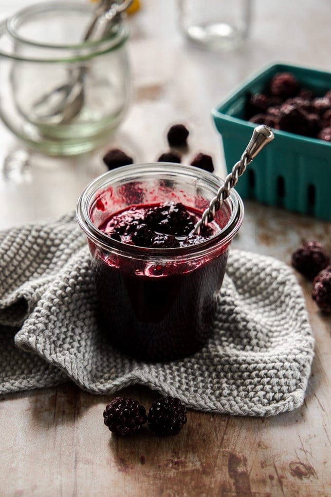 A jar of blackberry compote with a spoon sticking out sitting on a grey wash cloth.