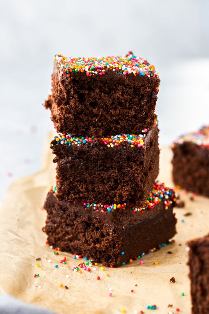 A stack of 3 square slices of chocolate cake topped with sprinkles stacked on a piece of brown parchment paper