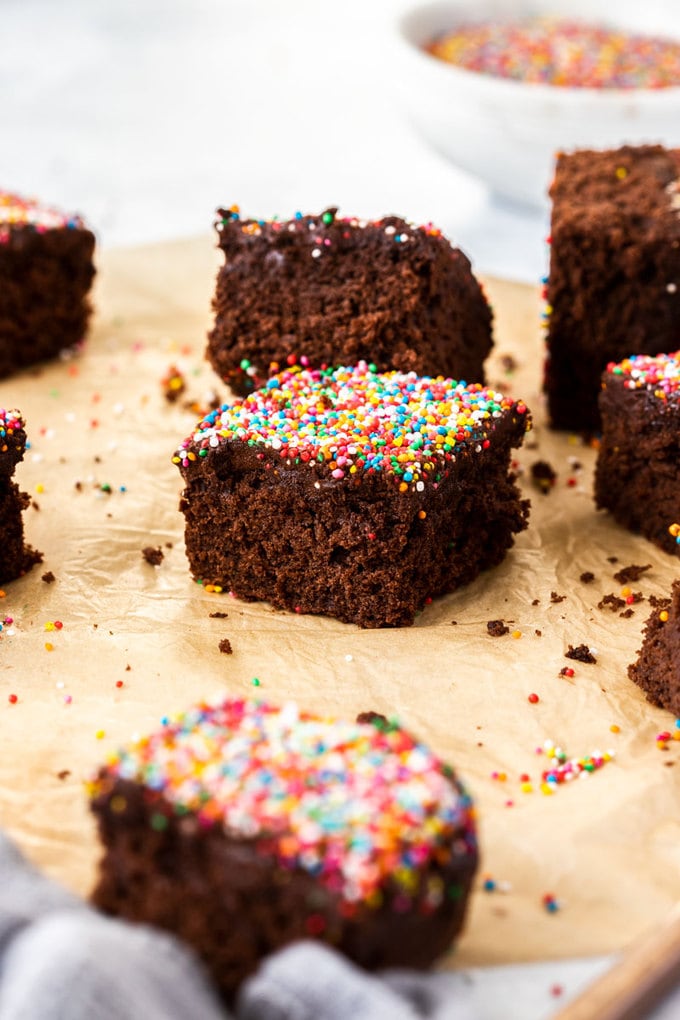 3 square slices of chocolate cake topped with sprinkles in lined up on a piece of brown parchment paper
