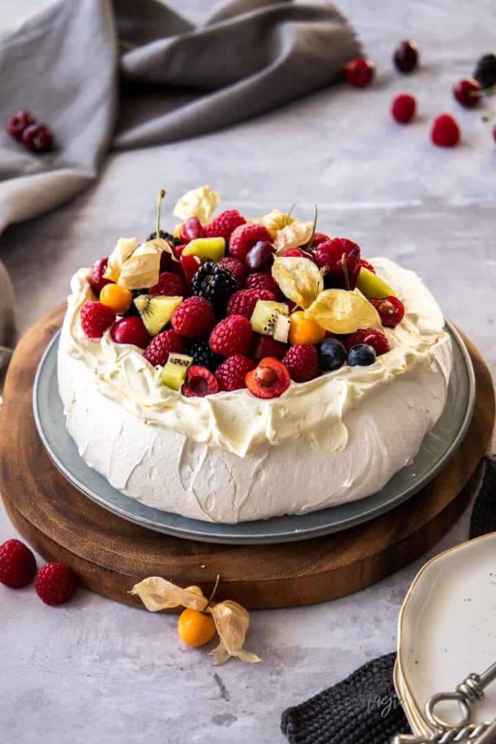 A pavlova topped with fresh berries and fruits on a wooden platter.