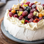 Close up of a pavlova topped with whipped cream and fruit on a wooden platter