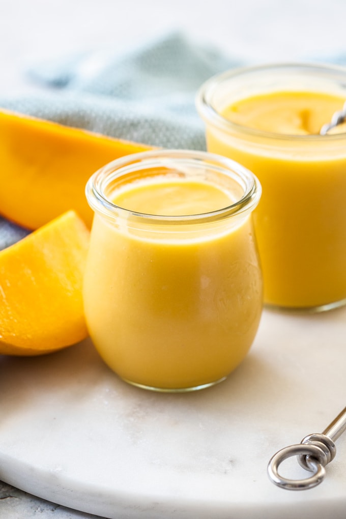 A small preserve jar filled with yellow mango curd, with another jar and slices of mango behind it.