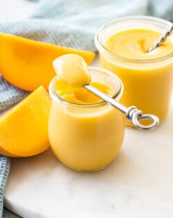 A small preserve jar filled with yellow mango curd, with another jar and slices of mango behind it.
