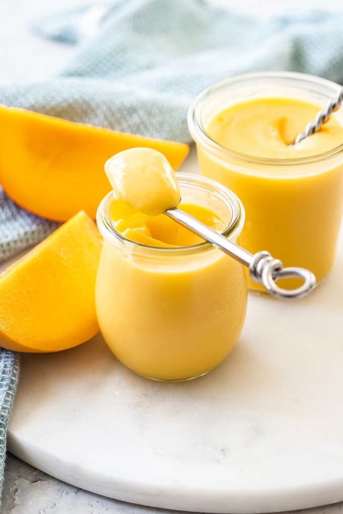 A spoon filled with mango curd resting on a small preserve jar filled with yellow mango curd, another jar and slices of mango behind it.
