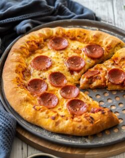 A pepperoni pizza on a metal tray on a wooden background. Once slice is cut out