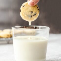 A glass of milk with a cookie being held above it and a drip of milk falling