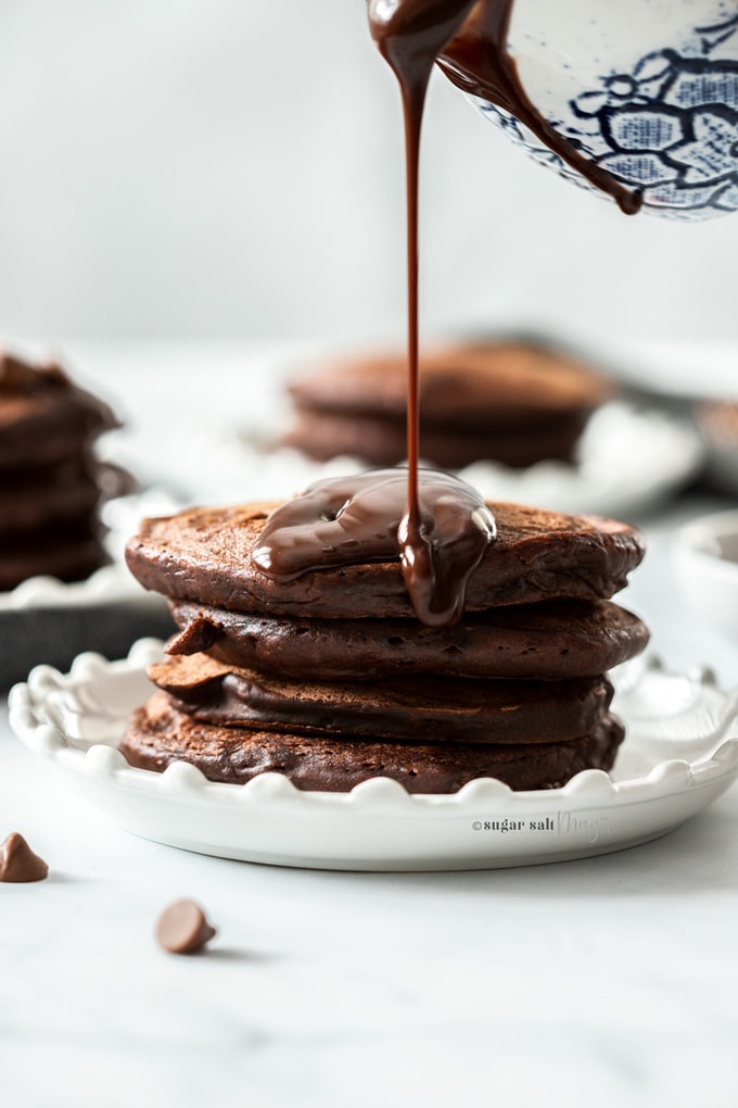 A stack of chocolate pancakes on a white plate with chocolate sauce being drizzled over the top