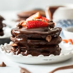 A stack of chocolate pancakes on a white plate, topped with strawberries chocolate chips and chocolate syrup.