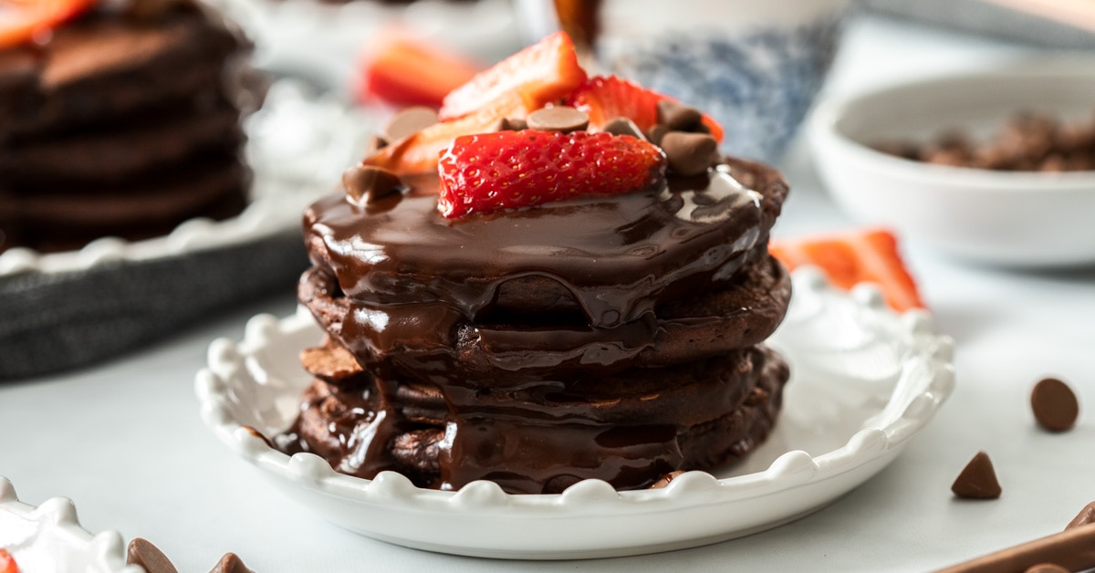A stack of chocolate pancakes on a white plate, topped with strawberries chocolate chips and chocolate syrup