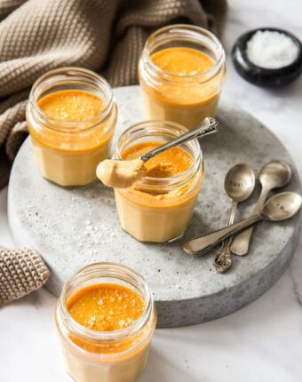 Small glass jars on a grey platter, filled with caramel custard. One has a spoon sitting on top