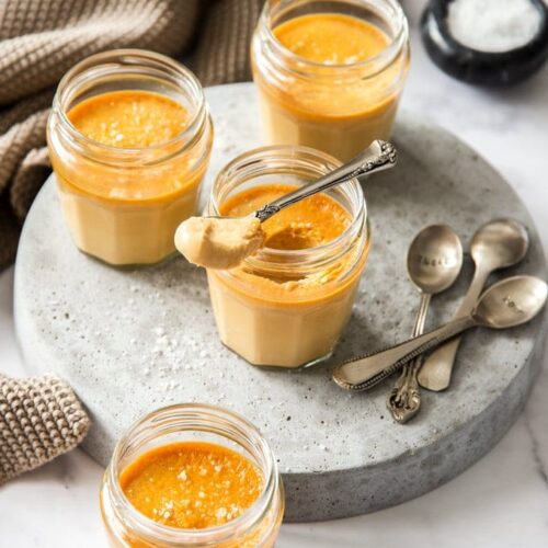 Small glass jars on a grey platter, filled with caramel custard. One has a spoon sitting on top