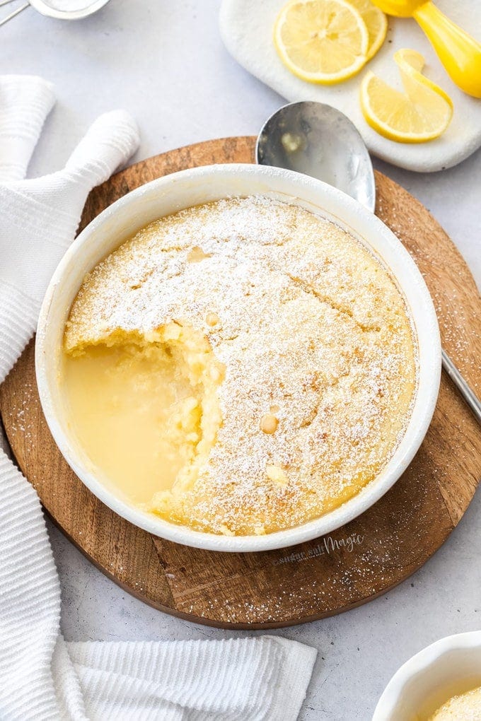 Top down view of a lemon pudding in a round white bowl, with a scoop taken out. Lemons in the top corner.