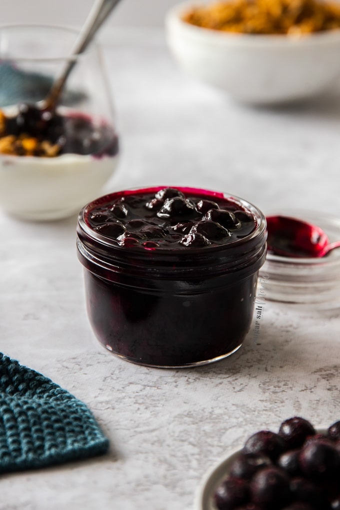 A small glass jar, filled to the brim with blueberry pie filling, yoghurt and granola in the background.