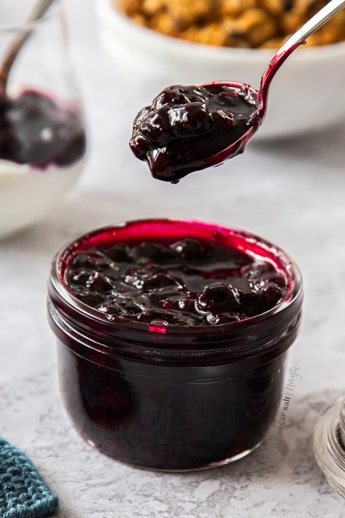 A small glass jar, filled to the brim with blueberry pie filling. A spoon filled with it hovers above.
