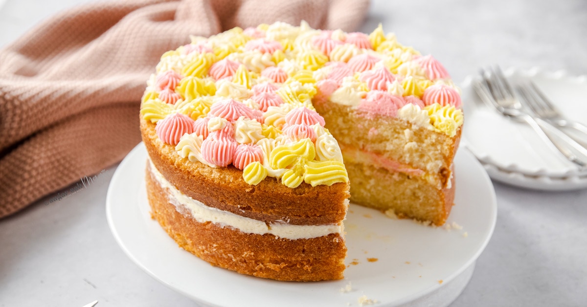 A vanilla cake with a slice remove, sitting on a white cake plate with a pink dish cloth in the background