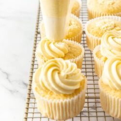 A row of cupcakes on a wire rack with frosting being piped on top