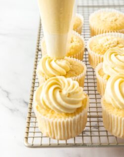 A row of cupcakes on a wire rack with frosting being piped on top