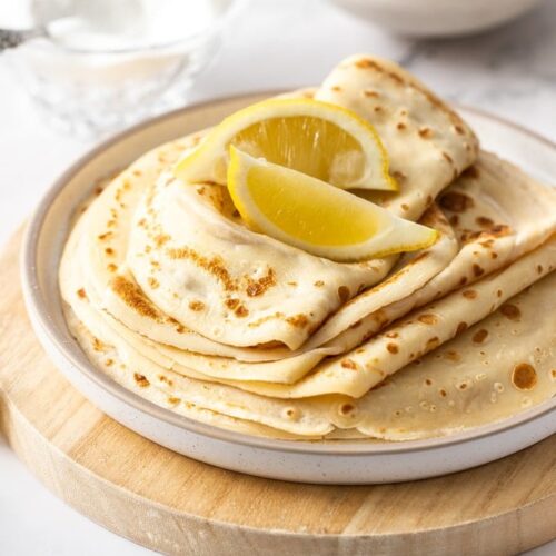 A white plate topped with a stack of english pancakes and wedges of lemon