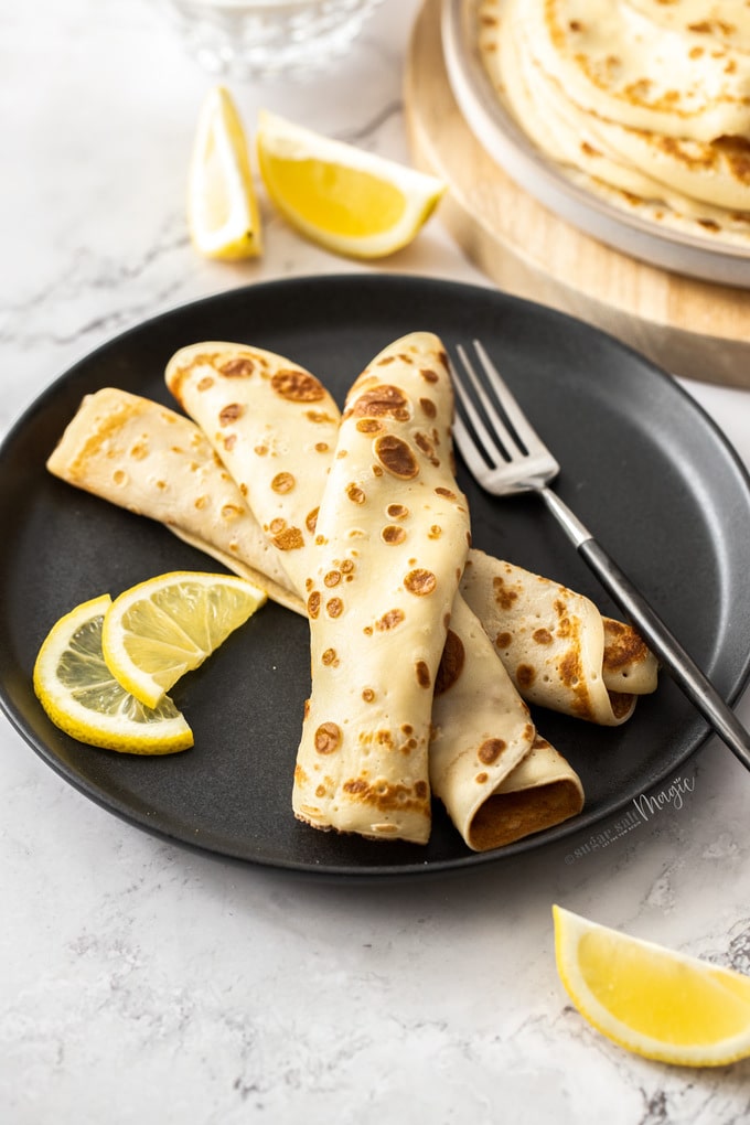 English pancakes rolled and stacked on a black plate with lemon wedges nearby