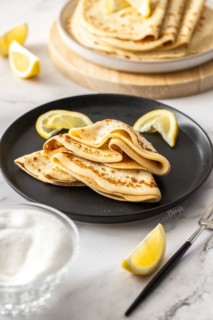 English pancakes folded and stacked on a black plate with lemon wedges nearby.