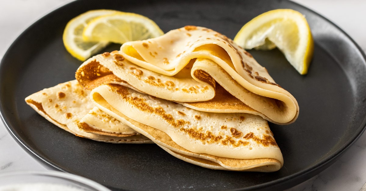 English pancakes folded and stacked on a black plate with lemon wedges nearby