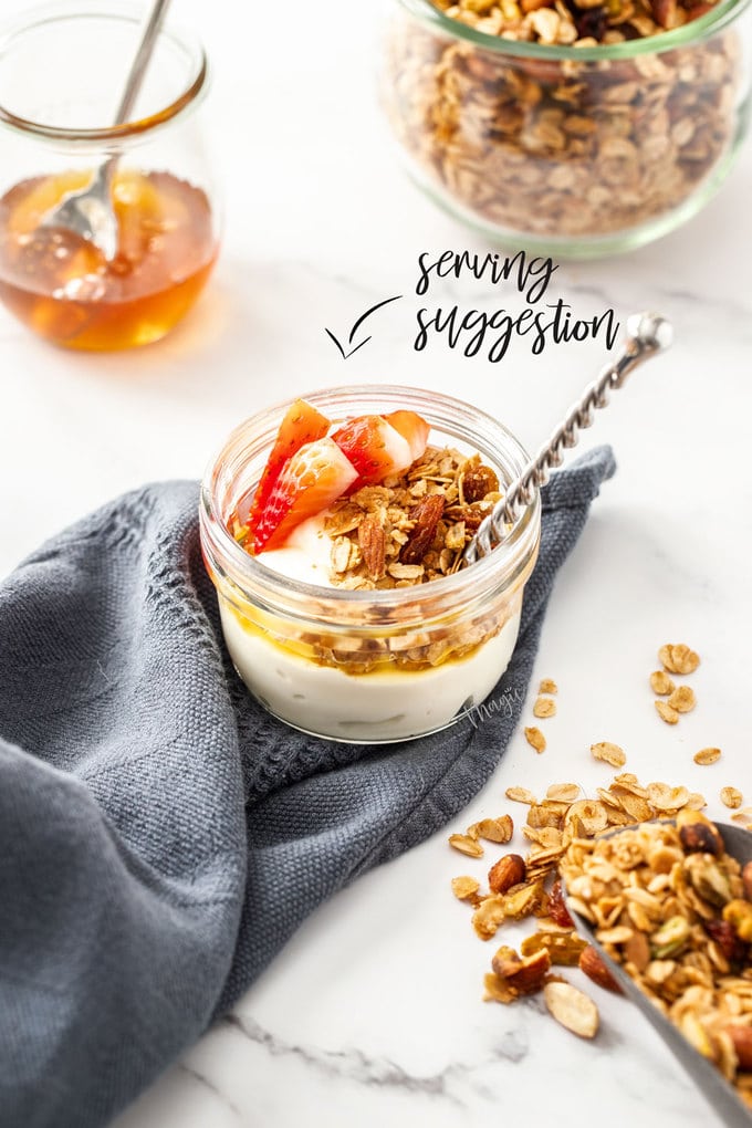 A small pot of yoghurt and granola. honey and more granola in the background