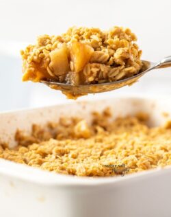 A spoon topped with apple crumble hovering above a dish full