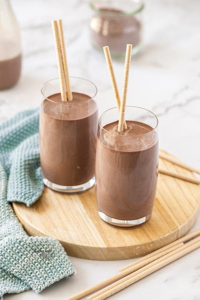 2 glasses of chocolate milk sitting on a small wooden platter