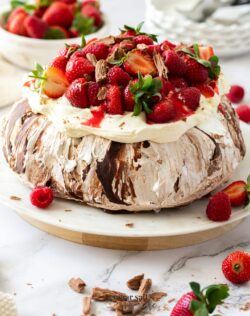 A chocolate pavlova topped with cream and berries on a white cake plate