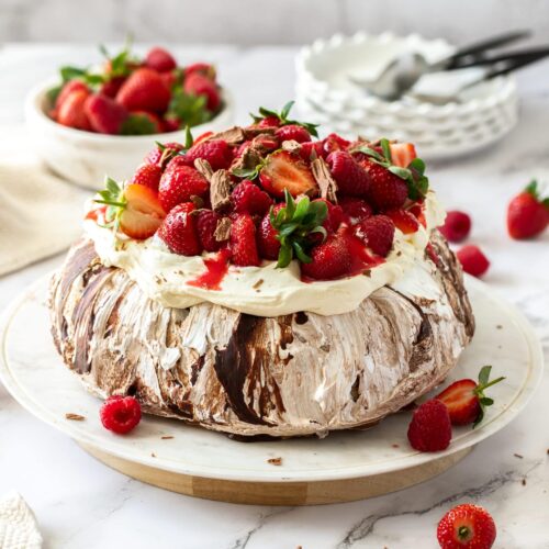 A chocolate pavlova topped with cream and berries on a white cake plate.