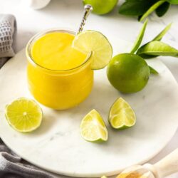 A glass jar filled with lime curd sitting on a white platter, surrounded by limes and eggs