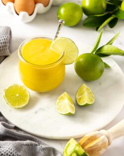 A glass jar filled with lime curd sitting on a white platter, surrounded by limes and eggs
