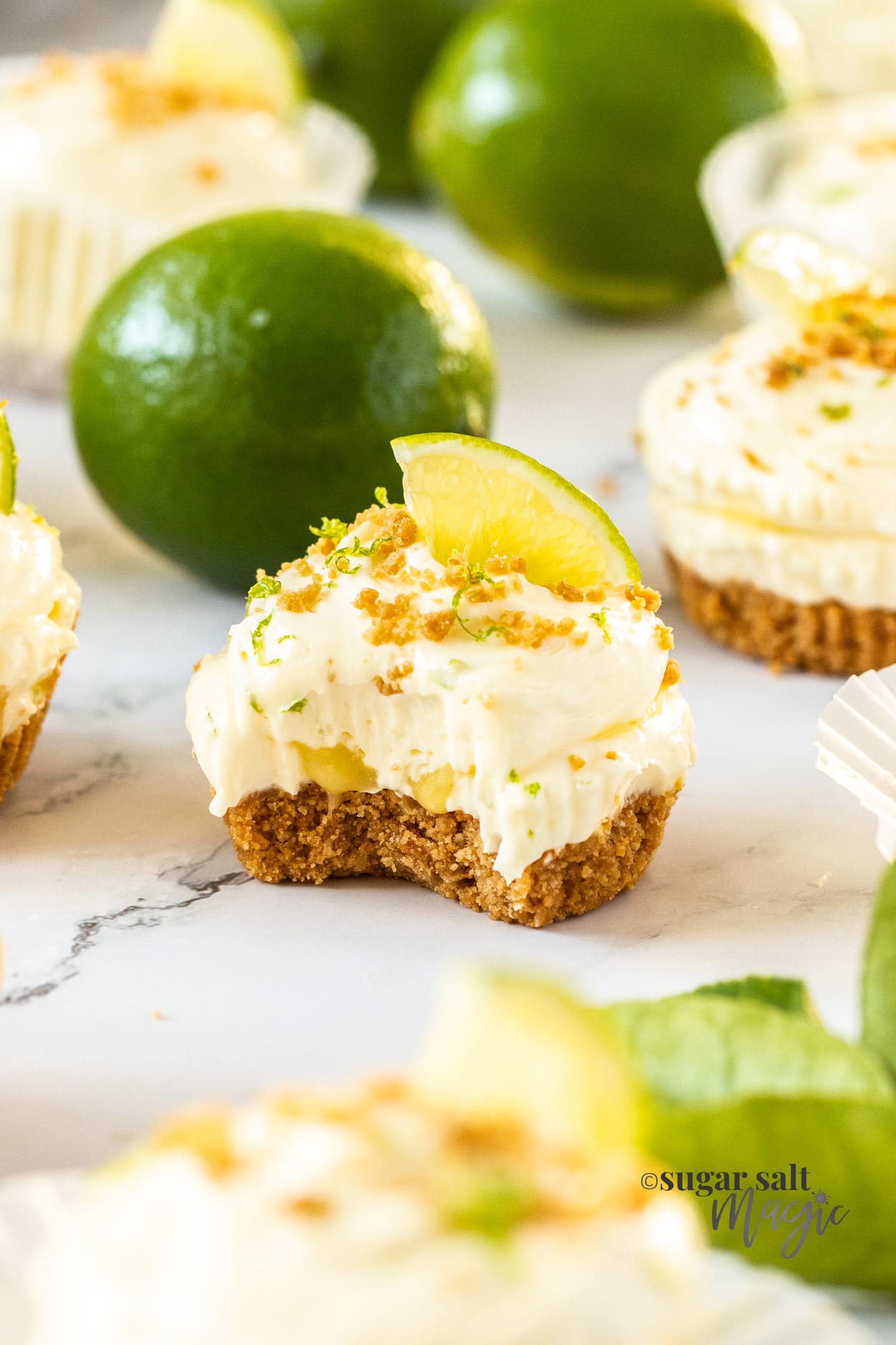 A mini lime cheesecake with a bite taken out and limes in the background