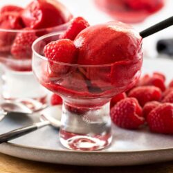 scoops of raspberry sorbet in a a small glass bowl with raspberries behind