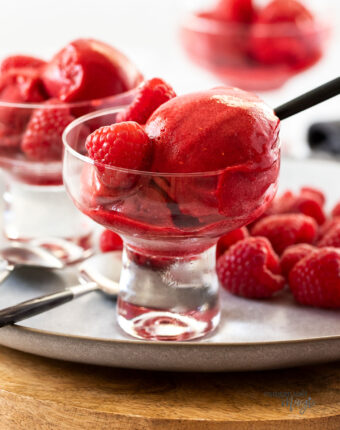 scoops of raspberry sorbet in a a small glass bowl with raspberries behind