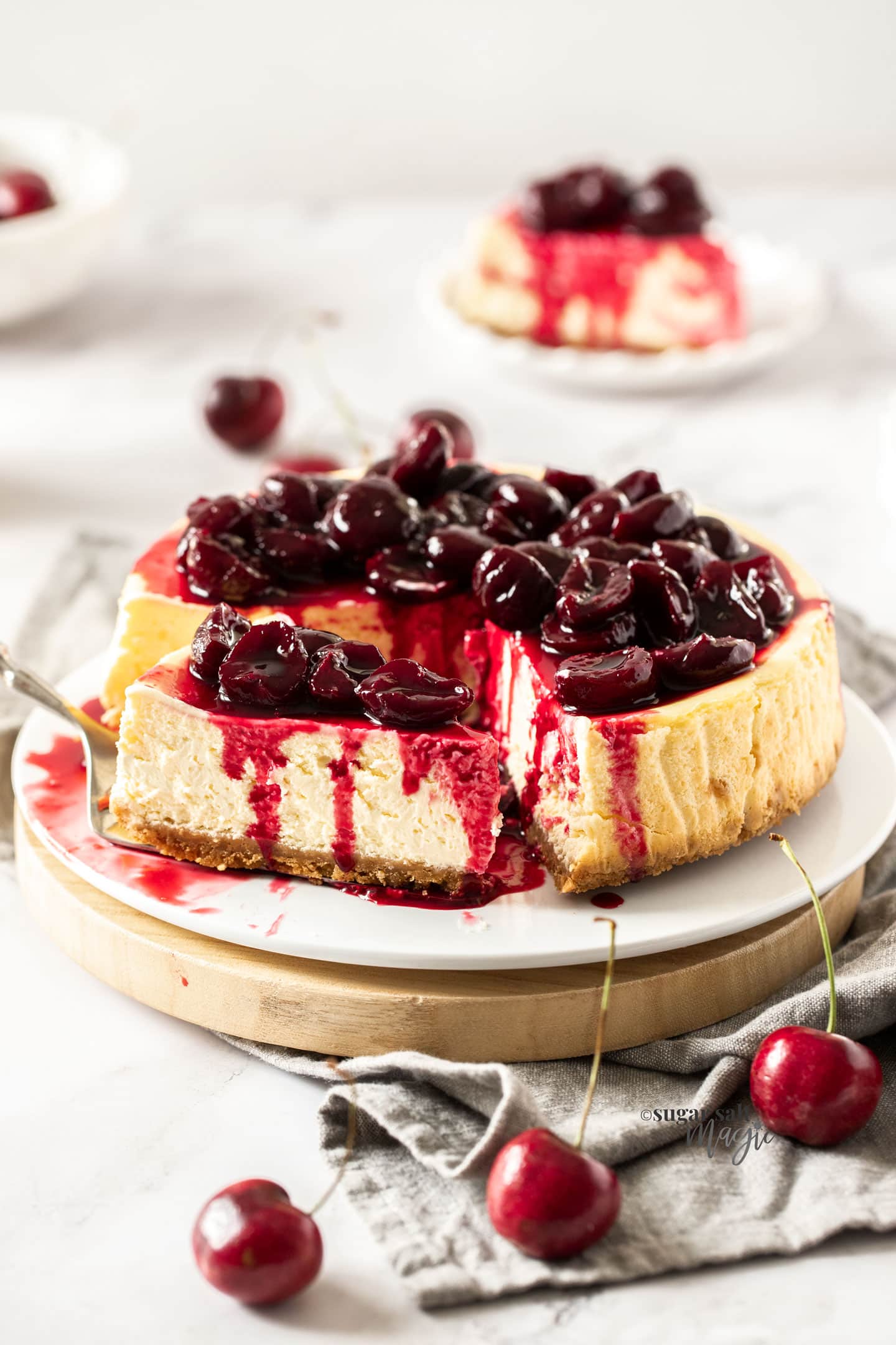 A cherry topped cheesecake on a white cake plaet with a slice in the background.