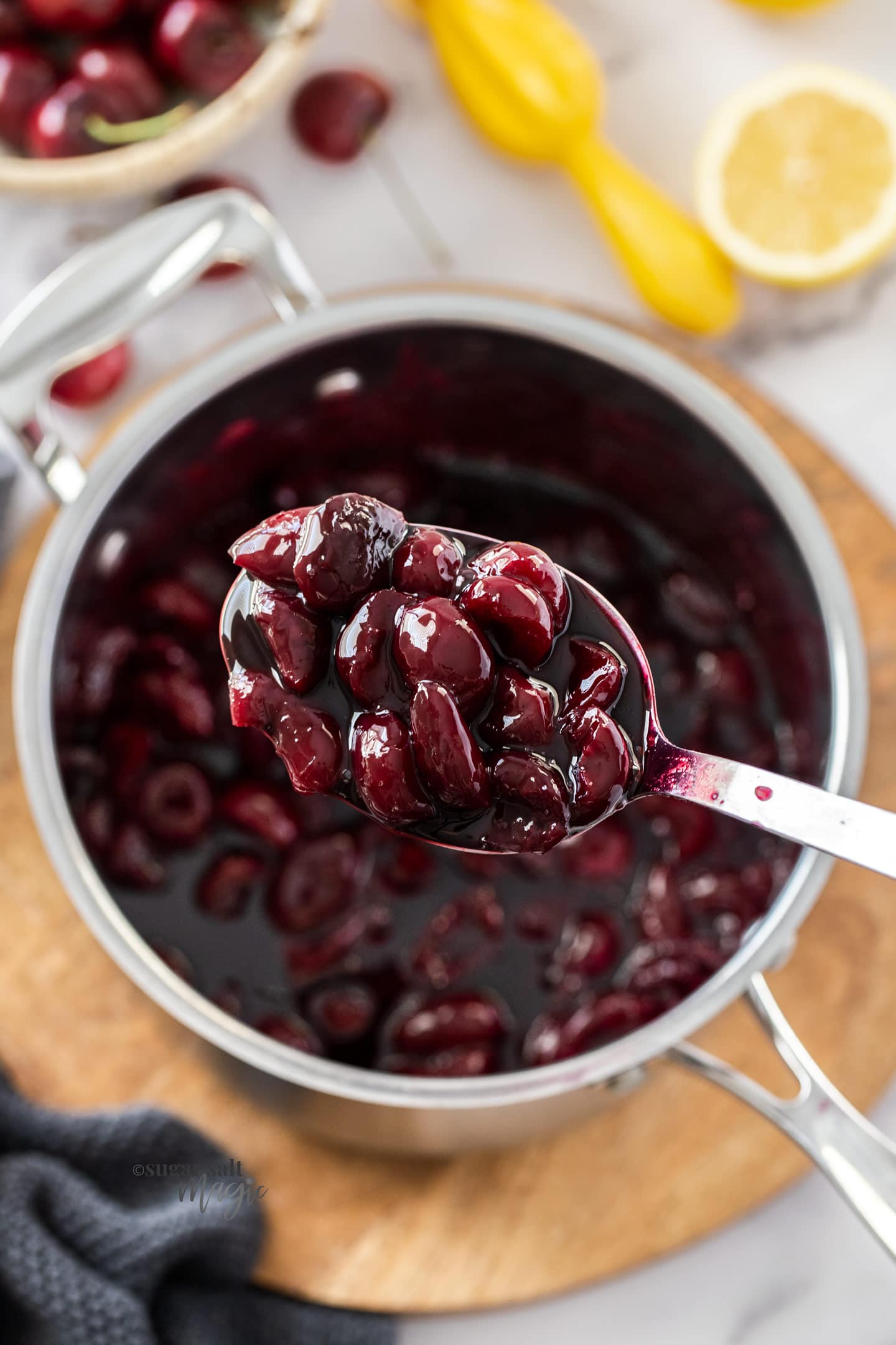 A spoon holding cherry sauce over a saucepan ful of it