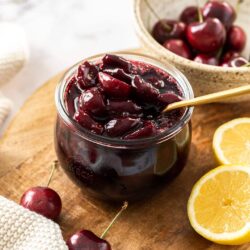 A glass jar filled with cherry compote with a gold spoon in it. Sitting on a wooden board.