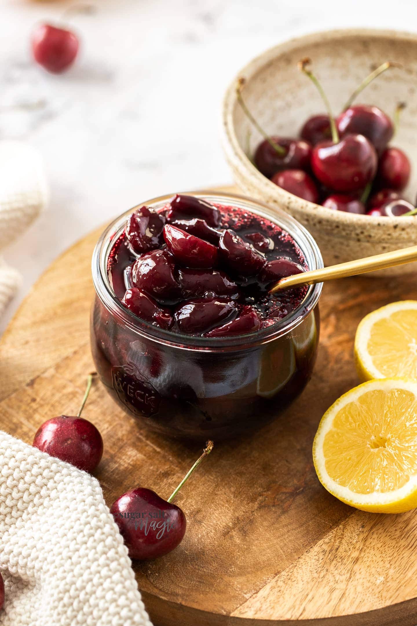 A glass jar filled with cherry compote with a gold spoon in it. Sitting on a wooden board