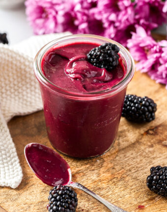 A glass jar filled with blackberry curd, surrounded by blackberries