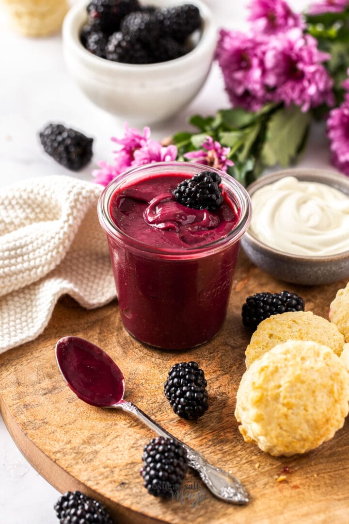 A glass jar filled with blackberry curd, surrounded by blackberries and scones
